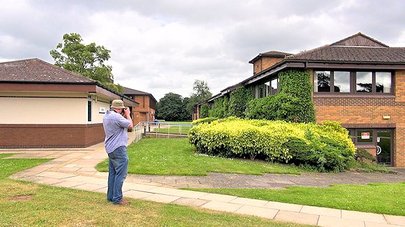 A man taking photographs with two buildings and shrubs behind
