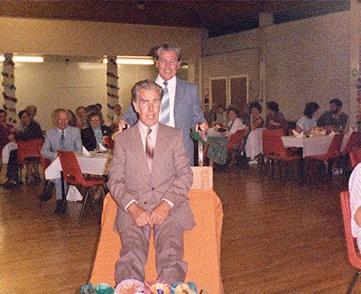 Chap in brown suit, sitting on a barrow, pushed by a chap in a grey-blue suit, in a canteen room, with people seated behind.